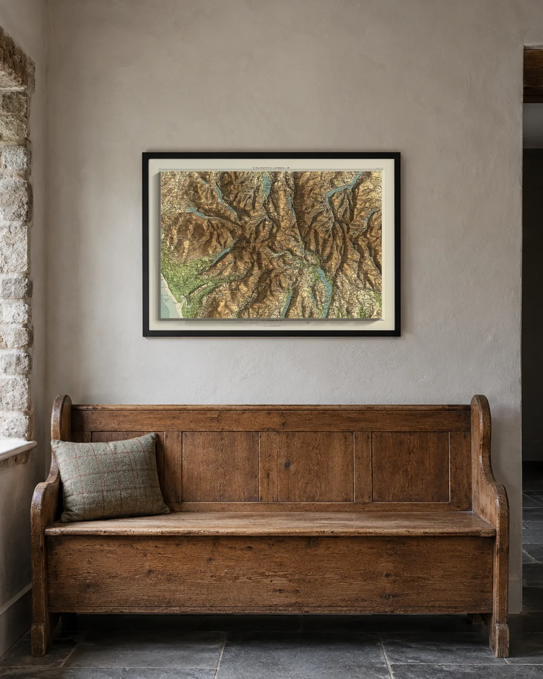 The Lake District Bartholomew edition, A1 in a black frame, hanging above a wooden settle in an illustrated stone cottage